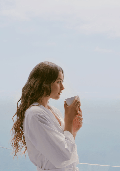 Woman in a white dress enjoying a cup of coffee on a balcony overlooking the sea